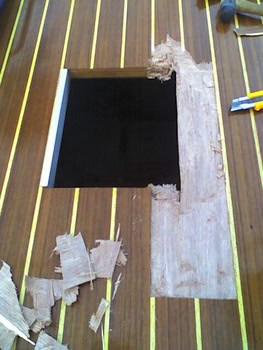 Un suelo de madera con un agujero rectangular, rodeado de trozos de madera arrancados y un cutter, que recuerda la precisión que requiere el minucioso mantenimiento de barcos en Barcelona de Grandval Náutico.