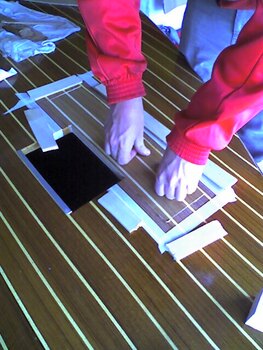 Una persona con una chaqueta roja prepara diligentemente un marco rectangular sobre una mesa de madera usando cinta blanca y papel, que recuerda la meticulosa artesanía que se ve en Grandval Náutico.