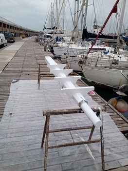 Un mástil de barco reposa horizontalmente sobre un muelle de madera, sostenido por caballetes, mientras varios yates están amarrados al fondo, un escenario ideal para que Grandval Náutico demuestre su experiencia en mantenimiento de barcos en Barcelona.