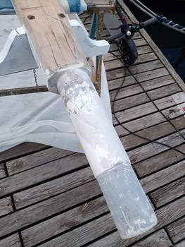 Un objeto cilíndrico de madera y metal, que recuerda a las herramientas esenciales para el mantenimiento de barcos en Barcelona, está parcialmente recubierto de pintura blanca y descansa sobre una cubierta de madera con elementos adicionales visibles en el fondo.