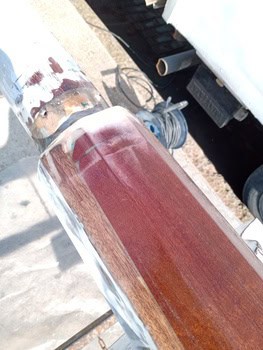 Primer plano de una barandilla de madera lijada durante un proceso de restauración en Grandval Náutico, con marcas visibles de lijado y algo de barniz eliminado. Al fondo, una bobina de cable y parte de un vehículo, escenas típicas del mantenimiento de barcos en Barcelona.