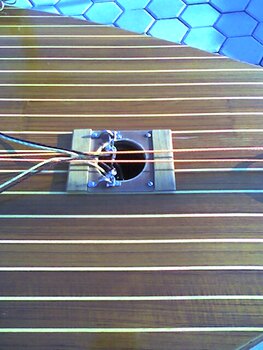 Un primer plano de un instrumento musical de cuerda con un cuerpo de madera y varias cuerdas dispuestas a través de un orificio central, similar a una cítara o un salterio, reposa sobre la superficie de baldosas como una obra de artesano en medio del bullicioso telón de fondo del Grandval Náutico.