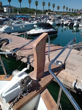 Un barco amarrado en el puerto deportivo Grandval Náutico muestra un poste de proa de madera, barandillas de metal y una cadena de ancla. Otros barcos y palmeras forman un pintoresco telón de fondo que resalta la vibrante escena de la vida marítima y el meticuloso mantenimiento de barcos en Barcelona.