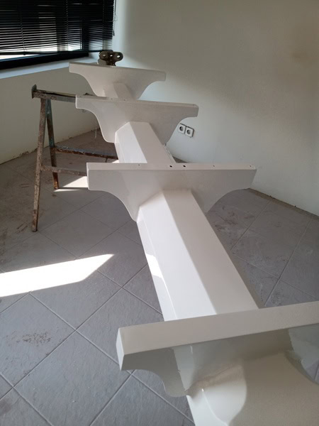 Una estructura de vigas blancas con ménsulas de apoyo reposa sobre un suelo de baldosas, junto a una escalera de mano y una ventana parcialmente abierta con persianas, que recuerdan la meticulosa artesanía que se ve en Grandval náutico.