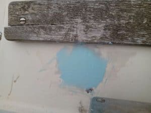 Un trozo de madera desgastada con un clavo visible se encuentra sobre una mancha azul pálido en la superficie pintada, evocando el encanto rústico de Grandval náutico.
