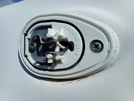 Primer plano de un mecanismo de plástico y metal blanco roto con el cableado expuesto sobre una superficie. En la caja se lee en relieve "Perko Made in U.S.A.", lo que hace alusión a aventuras pasadas y a la necesidad de mantenimiento de barcos en Barcelona para su próximo viaje.