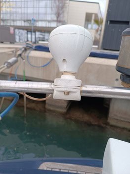 Una antena blanca instalada en un riel metálico cerca del agua en un puerto deportivo, con los edificios y equipos de Grandval Náutico visibles al fondo, mostrando el bullicioso centro de mantenimiento de barcos en Barcelona.