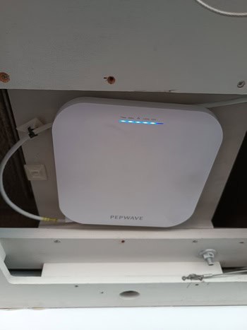 Un dispositivo Pepwave blanco, imprescindible para el mantenimiento de barcos en Barcelona, está montado en una pared. Sus luces indicadoras se iluminan en azul y hay varios cables conectados de forma experta.