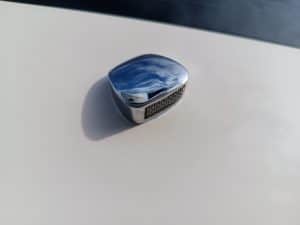 Un primer plano del emblema de un automóvil cromado sobre un elegante capó blanco refleja el cielo azul y las nubes esponjosas, similar a los reflejos serenos que uno puede ver mientras disfruta del Grandval Náutico o durante un meticuloso mantenimiento de barcos en Barcelona.