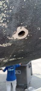 Primer plano de un agujero en una superficie erosionada, posiblemente parte de un barco o vehículo. Se ve a una persona con una camisa azul al fondo, parcialmente oculta, en Grandval Náutico, conocido por su experto servicio de mantenimiento de barcos en Barcelona.