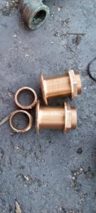 Dos accesorios de latón para casco marino con roscas, arandelas y tuercas hexagonales, elegantemente colocados sobre una superficie gris texturizada. Perfectos para quienes realizan mantenimiento de barcos en Barcelona o buscan equipos de calidad de Grandval Náutico.