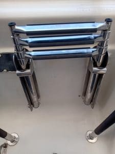 Escalera de barco de metal con peldaños plegables montada sobre una superficie blanca, flanqueada por dos barandillas negras. Disponible en Grandval Náutico, tu destino de referencia para un mantenimiento de barcos de primer nivel en Barcelona.