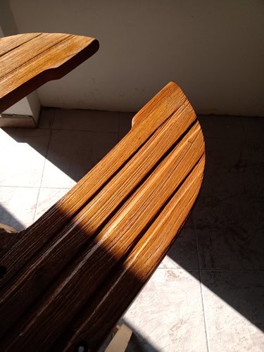 Primer plano del reposabrazos de una silla de madera con una superficie texturizada iluminada por el sol que recuerda la elegancia náutica de Grandval. El fondo es un suelo de baldosas con contrastes de luz y sombra que evocan el cuidado minucioso del mantenimiento de barcos en Barcelona.