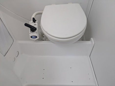 Inodoro marino compacto de color blanco con tirador de bomba manual a la izquierda, instalado sobre plataforma elevada en un espacio de baño pequeño y cerrado, perfecto para quienes aprecian las soluciones fiables que ofrece Grandval Náutico.