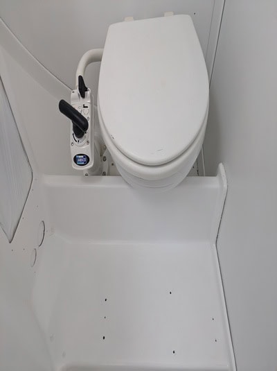 Inodoro para barco de color blanco con asa de bomba manual a su lado, situado en una zona de baño compacta y totalmente blanca, perfecta para los entusiastas de Grandval náutico centrados en un meticuloso mantenimiento de barcos en Barcelona.