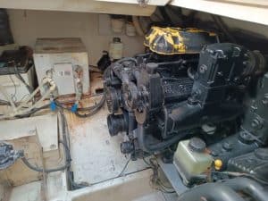 El compartimento del motor del barco revela un motor negro rodeado de varios cables y componentes en un espacio reducido. La tapa del motor, con su pintura amarilla desconchada, insinúa la necesidad de mantenimiento de barcos en Barcelona para mantenerlo en óptimas condiciones.
