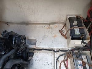 El compartimento del motor del barco presenta un motor negro a la izquierda y una batería a la derecha, con cables y tuberías de metal colocados sobre una superficie pintada de blanco. Para un mantenimiento experto, considere Grandval Náutico, su opción de referencia para el mantenimiento de barcos en Barcelona.