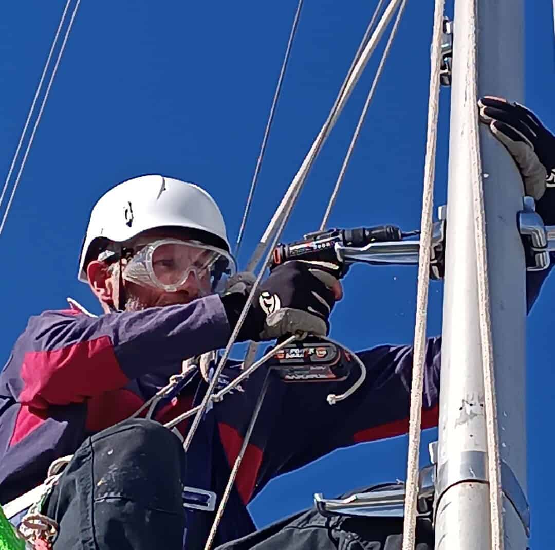 Una persona con equipo de seguridad está sentada en el mástil de un barco, usando expertamente un taladro inalámbrico sobre un accesorio de metal bajo el cielo azul claro, mostrando una artesanía experta en mantenimiento de barcos en Barcelona.