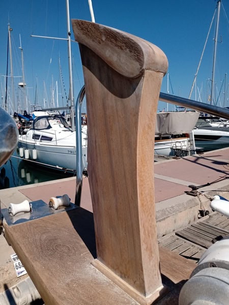 Un timón de barco de madera reposa en el muelle, rodeado de veleros bajo un cielo azul claro, reflejando la calidad artesanal de Grandval Náutico.