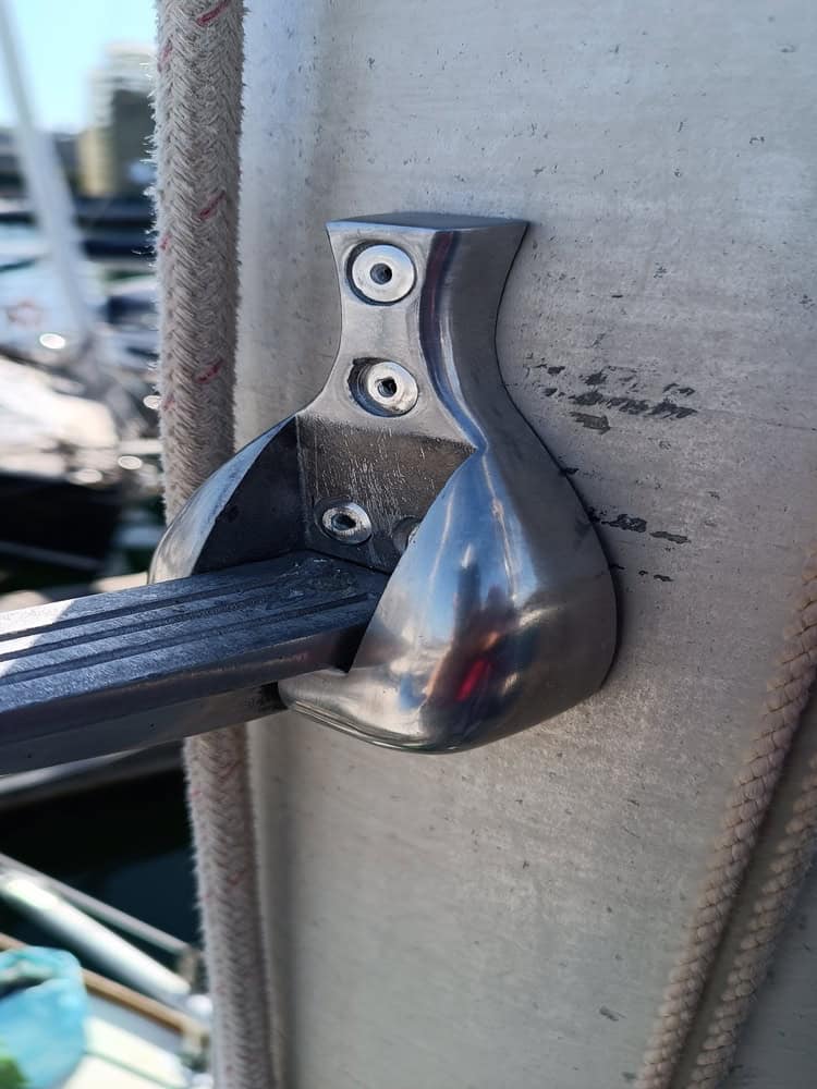Primer plano de un soporte metálico con tres tornillos que fijan una varilla rectangular a una superficie vertical, con un fondo borroso de cuerdas y barcos. Esta instantánea captura la esencia de Grandval náutico y su meticuloso mantenimiento de barcos en Barcelona.